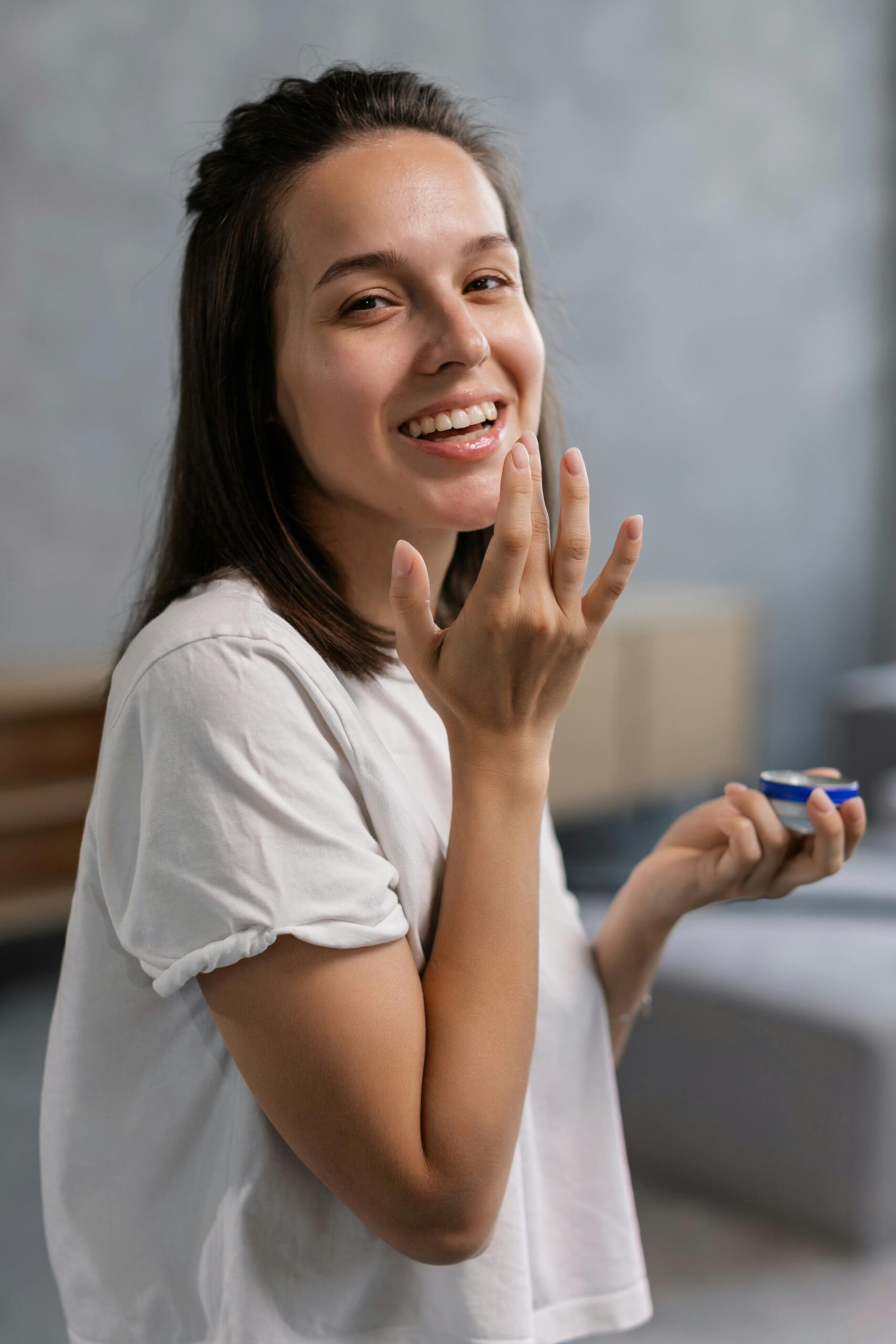 A cheerful woman applying moisturizing cream with a blurred background, indoors.